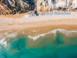 Aerial view of a sandy beach with clear blue water and gentle waves meeting the shore. Sunbeds and umbrellas are arranged neatly in rows, indicating a well-organized beach setup. Cliffs with vegetation rise at one end of the beach, adding a natural backdrop to the scene.