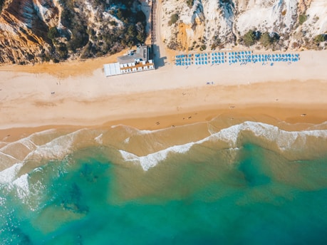 Aerial view of a sandy beach with clear blue water and gentle waves meeting the shore. Sunbeds and umbrellas are arranged neatly in rows, indicating a well-organized beach setup. Cliffs with vegetation rise at one end of the beach, adding a natural backdrop to the scene.