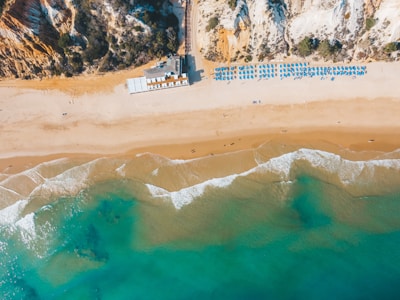 Aerial view of a sandy beach with clear blue water and gentle waves meeting the shore. Sunbeds and umbrellas are arranged neatly in rows, indicating a well-organized beach setup. Cliffs with vegetation rise at one end of the beach, adding a natural backdrop to the scene.