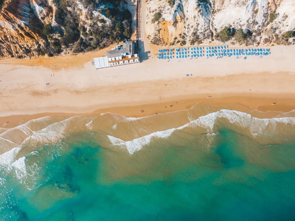 Aerial view of a sandy beach with clear blue water and gentle waves meeting the shore. Sunbeds and umbrellas are arranged neatly in rows, indicating a well-organized beach setup. Cliffs with vegetation rise at one end of the beach, adding a natural backdrop to the scene.