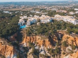 An aerial view of a coastal resort area featuring numerous white buildings with tan rooftops surrounded by lush greenery. There is a prominent cliff with orange and white rock formations, and dense forested areas. A swimming pool can be seen among the buildings, and there are further forested hills and structures in the distant background.