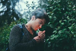 A person stands outdoors lighting a cigarette, surrounded by lush green foliage. The individual appears to be focused on their task, with a calm demeanor. The background is full of leafy plants, creating a natural and serene environment.