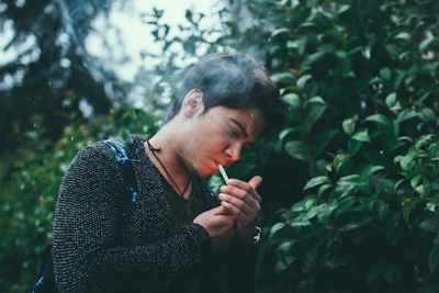 A person stands outdoors lighting a cigarette, surrounded by lush green foliage. The individual appears to be focused on their task, with a calm demeanor. The background is full of leafy plants, creating a natural and serene environment.