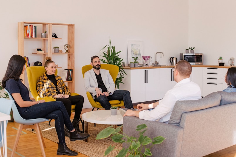 A group of people is seated in a casual office setting, engaged in conversation. The room features a modern design with a mix of furniture including armchairs and a sofa. A wooden bookshelf adorned with various items is placed against the wall, while a kitchenette is visible in the background. Several potted plants decorate the space, adding a touch of greenery.