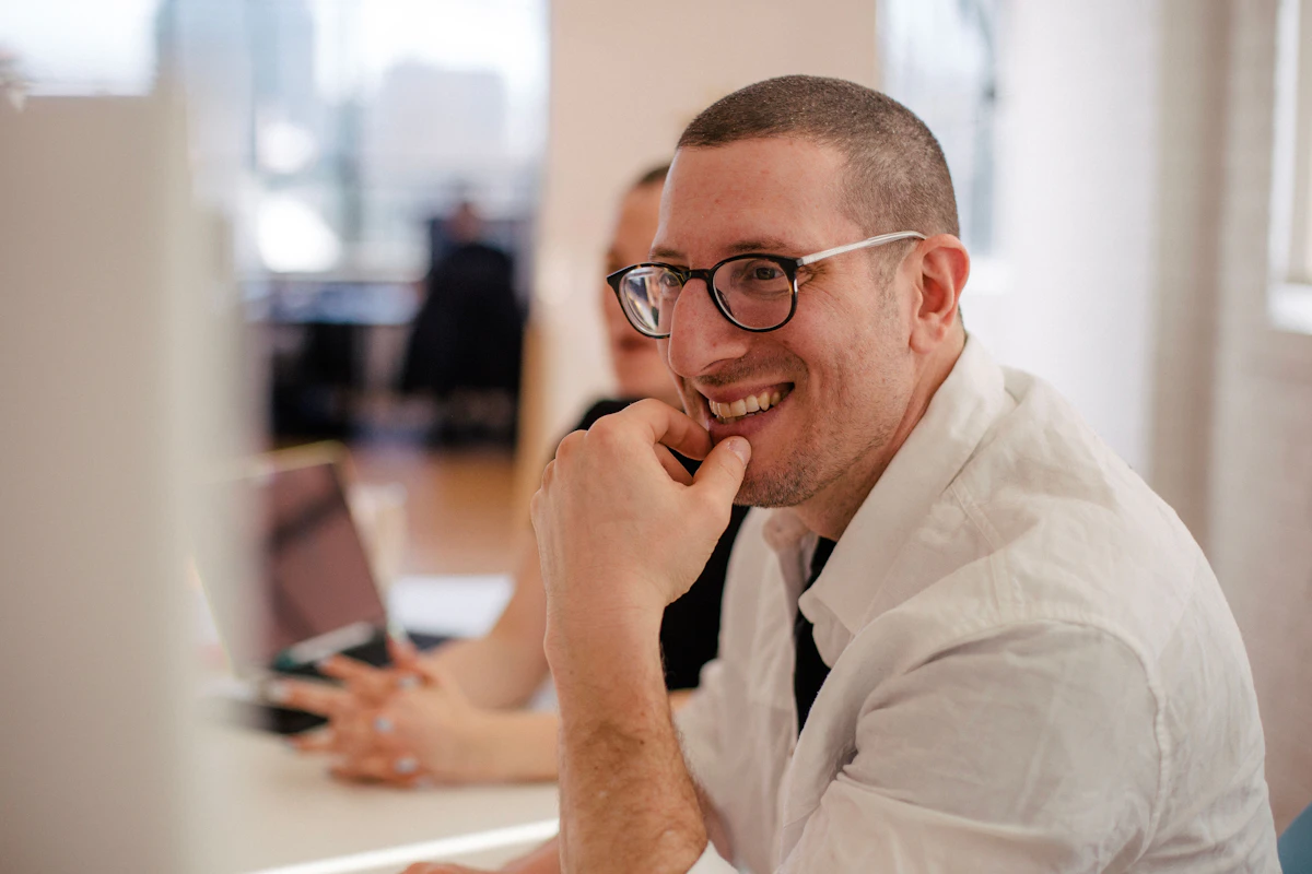 A man at his desk in a bright open-plan office, smiling as he looks up from his laptop