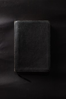 A closed book with a textured, dark cover is laid against a dark background. The book appears to have a ribbon bookmark.