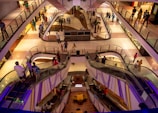 A modern shopping mall interior featuring multiple levels connected by escalators, with people walking and interacting on each level. The architecture is sleek and contemporary, incorporating glass and metal, while the lighting creates a warm ambiance. The area is bustling with shoppers, and there are small decorative elements like potted plants.