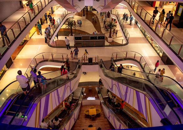 A modern shopping mall interior featuring multiple levels connected by escalators, with people walking and interacting on each level. The architecture is sleek and contemporary, incorporating glass and metal, while the lighting creates a warm ambiance. The area is bustling with shoppers, and there are small decorative elements like potted plants.