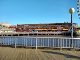 A waterfront scene includes a large brick and wooden building with signs reading 'Miners Landing'. The structure is adjacent to a pier with calm water, bordered by a metal fence. There is a ferris wheel partially visible on the right, and a clear blue sky above.