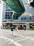 A modern shopping complex entrance with people walking. The architecture features a glass pedestrian bridge overhead and large glass windows. Various retail stores, such as Swatch and Aape, are visible with colorful signage.