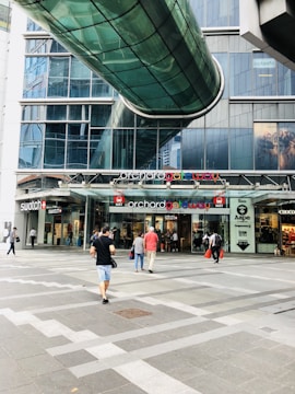 A modern shopping complex entrance with people walking. The architecture features a glass pedestrian bridge overhead and large glass windows. Various retail stores, such as Swatch and Aape, are visible with colorful signage.