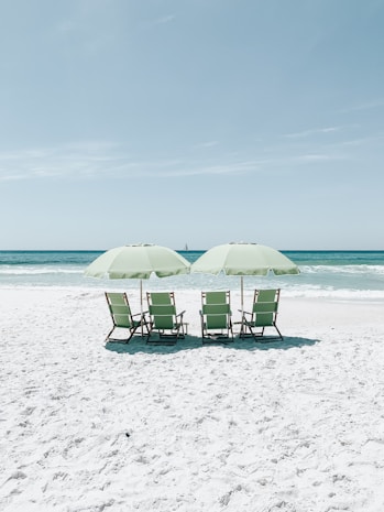 Four beach chairs under two large parasols are situated on a pristine white sandy beach facing the turquoise ocean. The sky above is clear with a pale blue hue, and there's a small sailboat in the distance on the water.