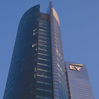 A modern high-rise building with a sleek glass facade and curved architecture against a clear blue sky. The structure features a prominent logo with the letters 'EY' on one of its sections.