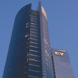 A modern high-rise building with a sleek glass facade and curved architecture against a clear blue sky. The structure features a prominent logo with the letters 'EY' on one of its sections.
