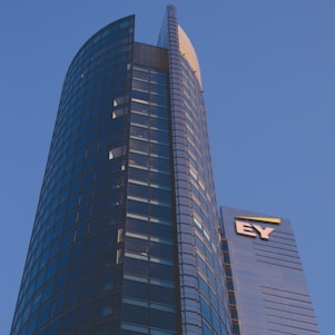 A modern high-rise building with a sleek glass facade and curved architecture against a clear blue sky. The structure features a prominent logo with the letters 'EY' on one of its sections.