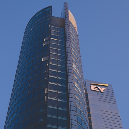 A modern high-rise building with a sleek glass facade and curved architecture against a clear blue sky. The structure features a prominent logo with the letters 'EY' on one of its sections.