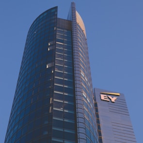 A modern high-rise building with a sleek glass facade and curved architecture against a clear blue sky. The structure features a prominent logo with the letters 'EY' on one of its sections.