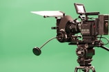 A professional video camera with a large lens is mounted on a tripod. The camera is equipped with various attachments and labeled 'RED DRAGON,' positioned against a plain green background.