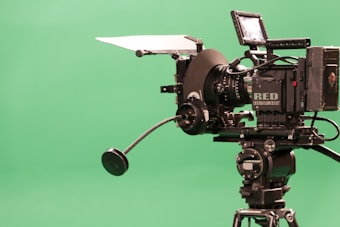A professional video camera with a large lens is mounted on a tripod. The camera is equipped with various attachments and labeled 'RED DRAGON,' positioned against a plain green background.