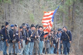A group of men dressed in historic military uniforms are standing together in a wooded area. They are holding rifles and an American flag with a combination of red, white, and blue horizontal stripes. The men are wearing dark blue jackets, light blue trousers, and hats.