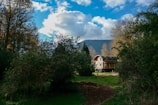 A wooden house with a curved roof surrounded by dense greenery and trees is set against the backdrop of a mountain range under a partly cloudy blue sky. A dirt path leads to the house, adding a rustic charm to the serene, natural landscape.