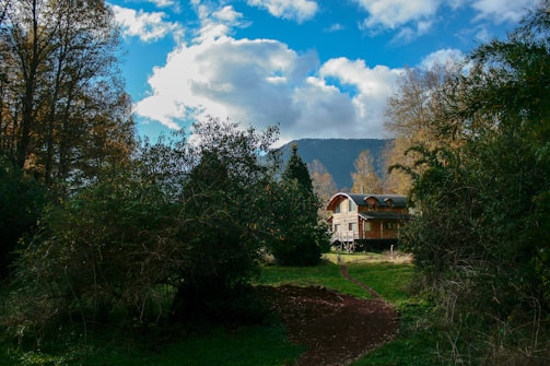 A wooden house with a curved roof surrounded by dense greenery and trees is set against the backdrop of a mountain range under a partly cloudy blue sky. A dirt path leads to the house, adding a rustic charm to the serene, natural landscape.