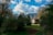 A wooden house with a curved roof surrounded by dense greenery and trees is set against the backdrop of a mountain range under a partly cloudy blue sky. A dirt path leads to the house, adding a rustic charm to the serene, natural landscape.