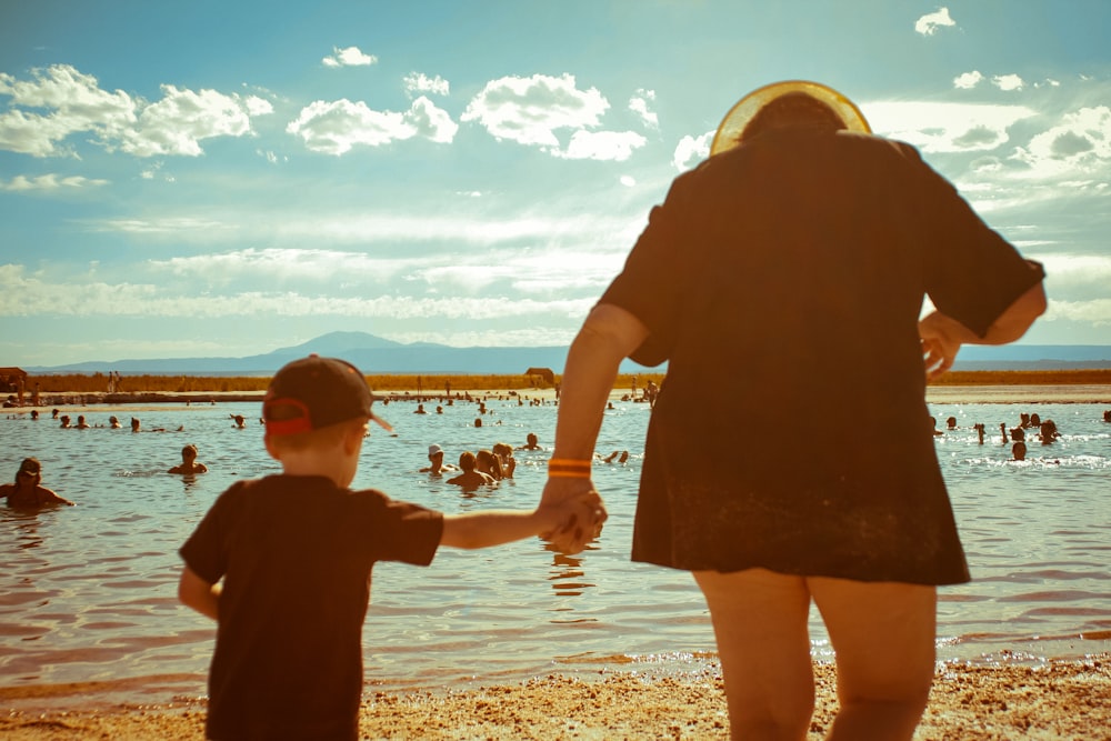 person holding hand of boy while walking near seashore and people swimming on sea under blue and white skies