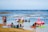 A group of people is at the beach, enjoying shallow waters with inflatable toys resembling a flamingo and a unicorn. Some are sitting on colorful beach chairs on the sand, while others are in the water, some of whom are in kayaks and inflatable floats.