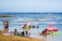 A group of people is at the beach, enjoying shallow waters with inflatable toys resembling a flamingo and a unicorn. Some are sitting on colorful beach chairs on the sand, while others are in the water, some of whom are in kayaks and inflatable floats.