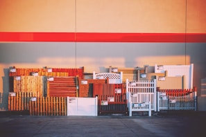 A variety of fence samples are displayed against a large wall, arranged in a row. The fences include different styles and materials such as wood and vinyl, in various colors such as brown, white, and red. Each fence section has a small sign attached, likely indicating pricing or specifications. The background features a wide red stripe across the wall, with shadows cast on the pavement below.