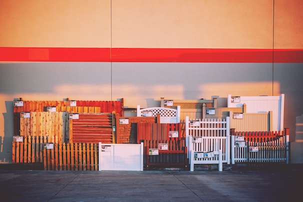 A variety of fence samples are displayed against a large wall, arranged in a row. The fences include different styles and materials such as wood and vinyl, in various colors such as brown, white, and red. Each fence section has a small sign attached, likely indicating pricing or specifications. The background features a wide red stripe across the wall, with shadows cast on the pavement below.