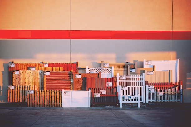 A variety of fence samples are displayed against a large wall, arranged in a row. The fences include different styles and materials such as wood and vinyl, in various colors such as brown, white, and red. Each fence section has a small sign attached, likely indicating pricing or specifications. The background features a wide red stripe across the wall, with shadows cast on the pavement below.