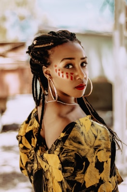 A person with braided hair and large hoop earrings gazes over their shoulder, wearing bold makeup with red patterns on the face. They are dressed in a striking garment with a yellow and black leaf pattern. The background is soft-focused, suggesting an outdoor setting with diffused natural light.