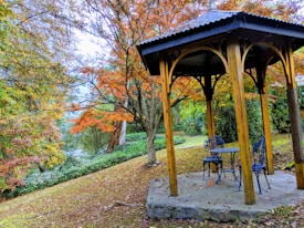 A wooden gazebo surrounded by lush greenery and vibrant autumn foliage. Two ornate blue metal chairs and a table are placed inside the structure, which is set on a concrete slab. The leaves on the trees display a mix of green, orange, and yellow hues, capturing the essence of a serene and picturesque garden in the fall season.