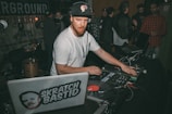 A DJ wearing a white t-shirt and cap is playing music on turntables in a lively indoor setting. Around him, people are gathered and chatting, with some musical instruments visible in the background. There are laptops and DJ equipment present, indicating a musical performance or party.
