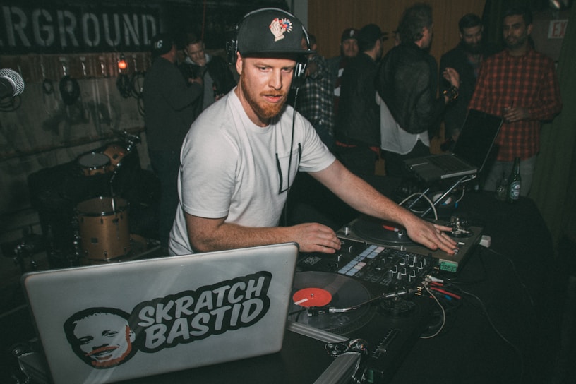 A DJ wearing a white t-shirt and cap is playing music on turntables in a lively indoor setting. Around him, people are gathered and chatting, with some musical instruments visible in the background. There are laptops and DJ equipment present, indicating a musical performance or party.