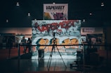 A dimly lit indoor scene with a large graffiti-covered wall serving as a backdrop. The graffiti is vibrant with colors like red and orange, featuring various tags and symbols. In front of the wall, a microphone stand is placed on a small stage or platform. There are several people in the background, possibly at an event or exhibition. Banners and signs are visible hanging above the scene, contributing to the industrial and urban atmosphere.