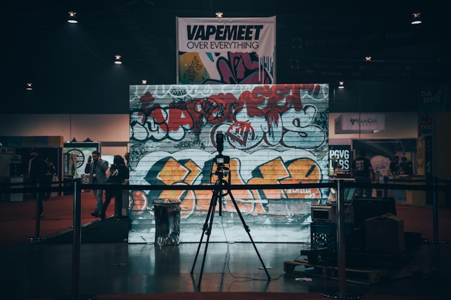 A dimly lit indoor scene with a large graffiti-covered wall serving as a backdrop. The graffiti is vibrant with colors like red and orange, featuring various tags and symbols. In front of the wall, a microphone stand is placed on a small stage or platform. There are several people in the background, possibly at an event or exhibition. Banners and signs are visible hanging above the scene, contributing to the industrial and urban atmosphere.