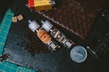 Two bottles of e-liquid labeled 'Creme De La Creme' placed on a surface with coffee beans scattered around them. Nearby are various items including a woven leather pouch, a Kodak film box, camera lens parts, and a cutting mat with grid lines.