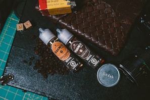 Two bottles of e-liquid labeled 'Creme De La Creme' placed on a surface with coffee beans scattered around them. Nearby are various items including a woven leather pouch, a Kodak film box, camera lens parts, and a cutting mat with grid lines.