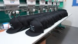A row of identical black caps is neatly lined up on a white table in front of a large embroidery machine, with threads and machinery parts visible. The caps feature a white embroidered design or logo on the front.