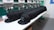 A row of identical black caps is neatly lined up on a white table in front of a large embroidery machine, with threads and machinery parts visible. The caps feature a white embroidered design or logo on the front.