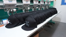 A row of identical black caps is neatly lined up on a white table in front of a large embroidery machine, with threads and machinery parts visible. The caps feature a white embroidered design or logo on the front.