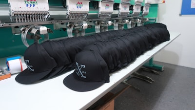 A row of identical black caps is neatly lined up on a white table in front of a large embroidery machine, with threads and machinery parts visible. The caps feature a white embroidered design or logo on the front.
