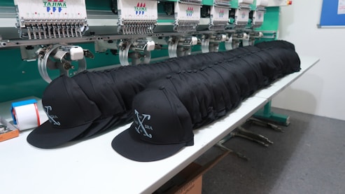 A row of identical black caps is neatly lined up on a white table in front of a large embroidery machine, with threads and machinery parts visible. The caps feature a white embroidered design or logo on the front.