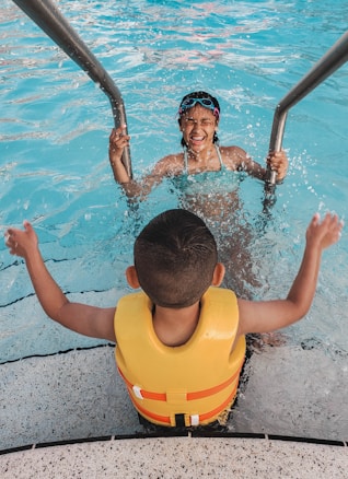 Two children are at a swimming pool. One child with goggles is climbing up the pool ladder, smiling joyfully, while another child is sitting at the edge of the pool, wearing a yellow life jacket. The water in the pool is a clear blue, and both children appear to be having fun.