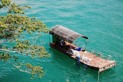 A small, rustic boat with a canopy floats on clear turquoise water. An assortment of items, including blue barrels and white containers, are visible on the boat deck. The boat is tied to the shore, and leafy tree branches extend into the frame from the left.