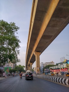 An elevated metro rail track runs parallel to a busy urban road. Vehicles, including a black car and a motorbike, are traveling under the track. Buildings with colorful signs line the road, and a large tree is present on the left side of the image. The sky is a clear light blue, indicating daytime.