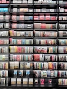 A large bookshelf filled with a variety of books stacked neatly in rows. The books have colorful spines with a mix of titles and authors visible. The shelf is black, creating a contrast with the diverse colors of the books.